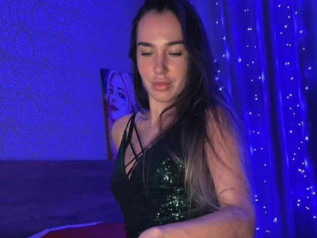 AraiaSheri with long brown hair, green sequined top, and tattoo on right arm, looking down with a slight smile in blue-lit room. Boobs visible under top.