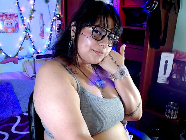 LilithHeavy with black hair, glasses, and nose ring, wearing a gray sports bra, showing her tits, smiling, hand on cheek, tattoo on wrist, in a colorful room.
