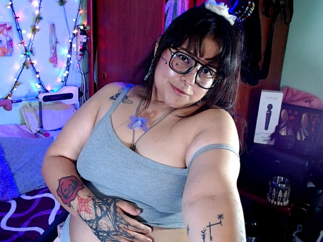 Lilith: Uma mulher com óculos, camiseta cinza, tatuagens, piercing no nariz e cabelo escuro, segurando um celular, mostrando os seios, tatuagem no braço direito, mão esquerda na barriga, sorrindo. Quarto, com luzes coloridas..