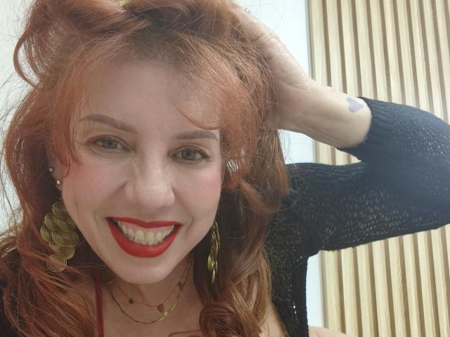 Khharha with red hair, black cardigan, red lipstick, gold earrings, smiling, hand in hair, visible tattoo on arm, wearing necklace, looking at camera, cute, attractive, sexy face.