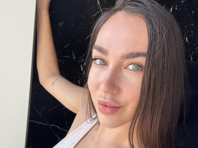 AraiaSheri has green eyes, straight brown hair, and a small tattoo on her upper arm. She's wearing a pink tank top, standing with her arm against a black wall.
