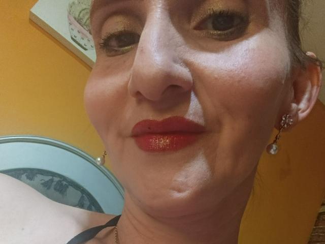 RubiAnderson has shiny red lipstick, gold eyeshadow, and a slight smile. She's wearing a black top, pearl earrings, and her face shows some wrinkles and subtle makeup.