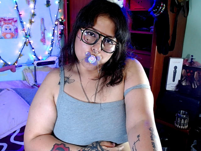 Lilith, con gafas, cabello negro y un chupete en la boca, vestida con una camiseta gris, mostrando tatuajes en sus brazos, y sosteniendo sus pechos en un dormitorio..