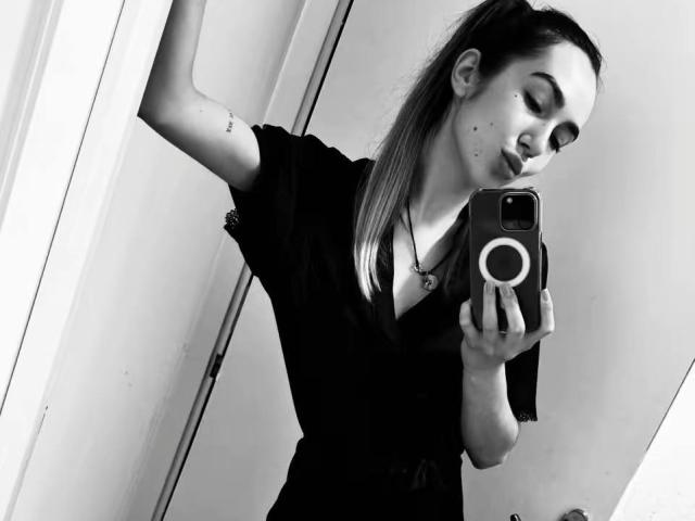 Selfie em preto e branco de AraiaSheri com um rabo de cavalo comprido, vestindo uma camisa escura, segurando um celular com uma capinha, mostrando uma tatuagem no braço superior e fazendo um beicinho..