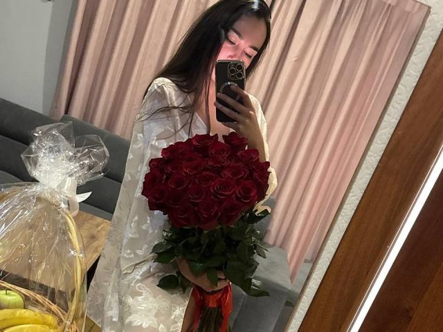 AraiaSheri, com cabelos castanhos longos, segurando um grande buquê de rosas vermelhas, vestindo um vestido branco transparente, tirando uma selfie no espelho, cortinas rosa ao fundo, bananas em uma cesta..