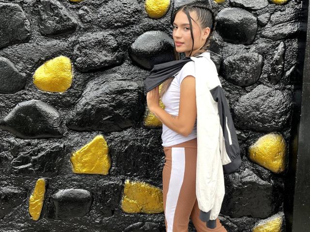 MillyPalmer with small tits, brown and white leggings, white and black top, black and white backpack, standing against black and yellow rock wall, looking over shoulder, smiling.