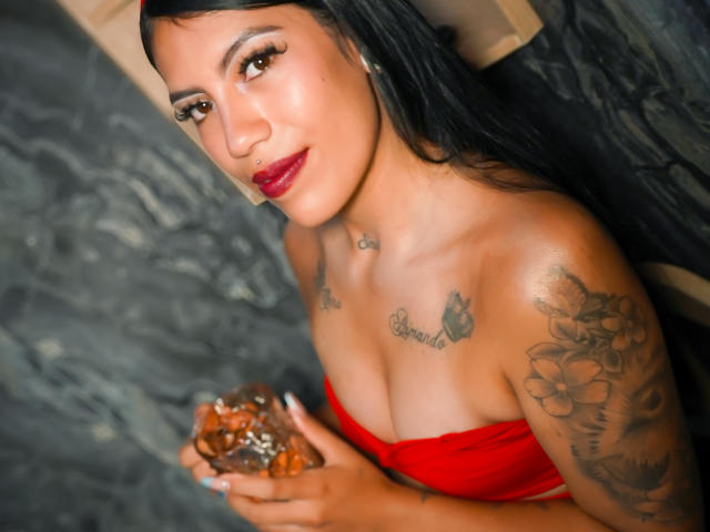 KymCorzo, con cabello oscuro, vestido rojo, tatuajes, sosteniendo caramelos, mirando a la cámara, sonriendo, con lápiz labial rojo, un pequeño aro en la nariz, piel oscura, senos de tamaño medio, tatuajes en los brazos, uñas negras..