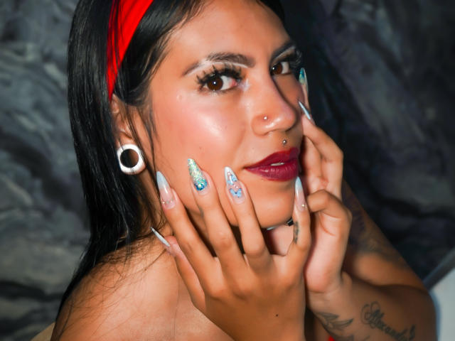 KymCorzo with dark hair, red headband, nose ring, large white ear gauges, long nails, colorful nail art, and tattoos, smiling, hands on face, wearing red lipstick.