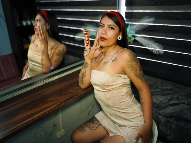 KymCorzo, avec des tatouages, portant un bandeau rouge et une robe courte beige, tenant un bonbon rayé, assise près d'un miroir qui la reflète. Des tatouages visibles sur les seins, les fesses et la vulve..