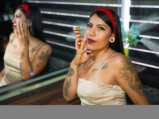 KymCorzo, avec ses cheveux foncés, un bandeau rouge, une robe blanche en dentelle, des tatouages, de gros piercings aux oreilles, tenant des bonbons, un miroir réfléchissant, une table en bois, du rouge à lèvres, regardant la caméra, montrant ses seins..