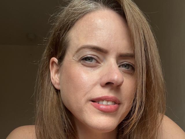 ElizabethArdente has light brown, shoulder-length hair, green eyes, and pink lips. Her face is slightly flushed with visible pores. She's looking at the camera with a subtle smile.