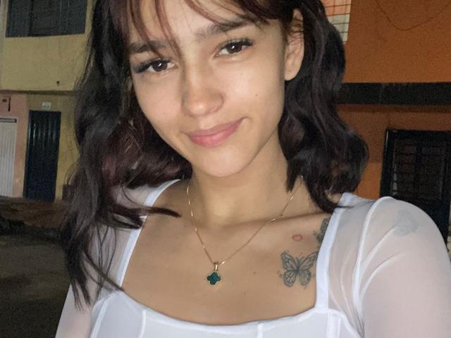MelodyMansson with dark wavy hair, smiling, wearing a white sheer top, necklace with green gem, tattooed butterflies on chest, brown eyes, and light skin.