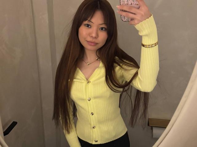 Joetta Bealer mit langen braunen Haaren, einem gelben Pullover und schwarzen Hosen, macht ein Selfie vor einem Spiegel und zeigt ihre kleinen Brüste und ihr hübsches Gesicht. Sie trägt ein goldenes Armband und eine Halskette..