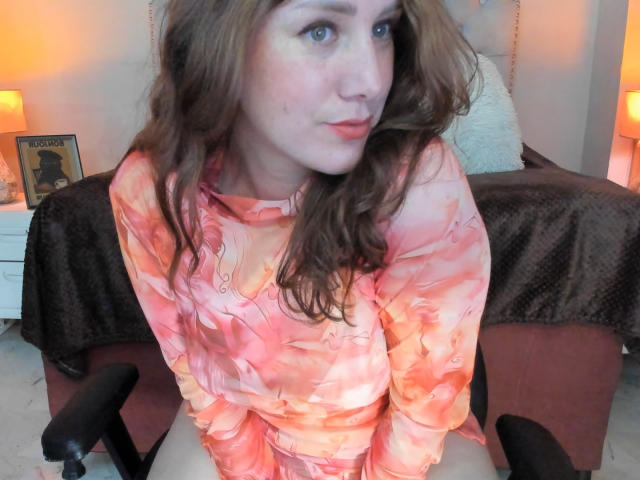 KatyaRogersX with wavy brown hair, pale skin, and blue eyes, wearing an orange and pink floral dress, sitting on a black chair, looking slightly to the side.