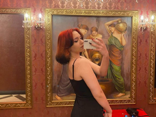DinahLackey with red hair, black dress, and visible ass, taking a mirror selfie in a fancy room, standing in front of a painting with classical figures.