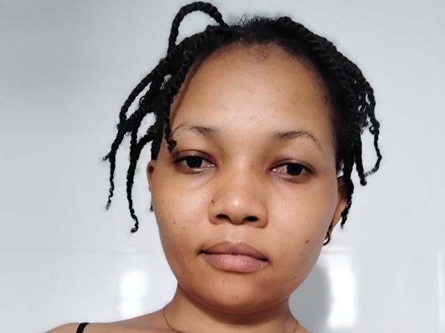 NessieCecily hat eine mittelbraune Hautfarbe, einen kleinen Nasenring und kurze, schwarze Dreadlocks. Sie blickt direkt in die Kamera mit einem neutralen Gesichtsausdruck und trägt ein schwarzes Oberteil..