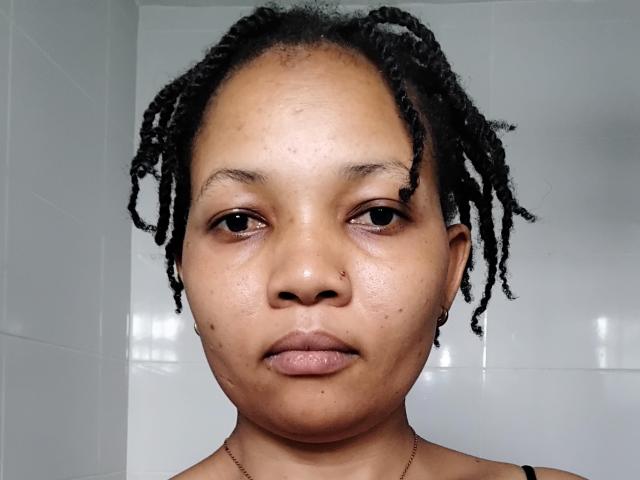 NessieCecily hat eine dunkelbraune Haut, eine kleine Nasenpiercing und kurze, schwarze Dreadlocks. Ihr Gesicht zeigt einen neutralen Ausdruck mit leicht geöffneten, rosa Lippen. Sie trägt eine dünne Halskette..