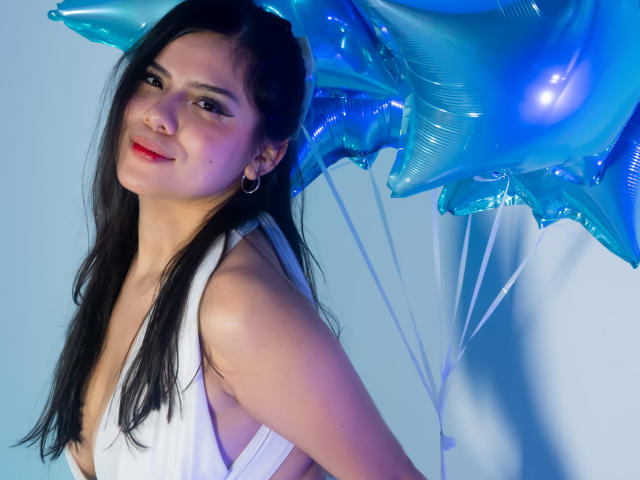 MaraCortes with long black hair, white dress, red lipstick, holding blue star balloons, smiling, wearing small hoop earrings, light blue background, soft blue and purple lighting.