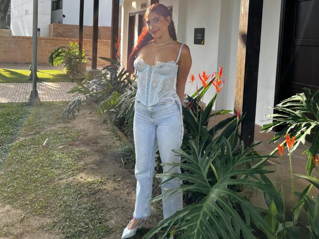 MiaBianchi with long red hair, wearing a lace-up blue corset and light blue jeans, standing by orange bird-of-paradise flowers, hands in pockets, white flats, sunny day.
