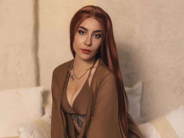 AlesziaRose with long red hair, brown eyes, and red lipstick, wearing a brown bikini top and sheer brown robe, showing her small tits and a tattoo on her chest.