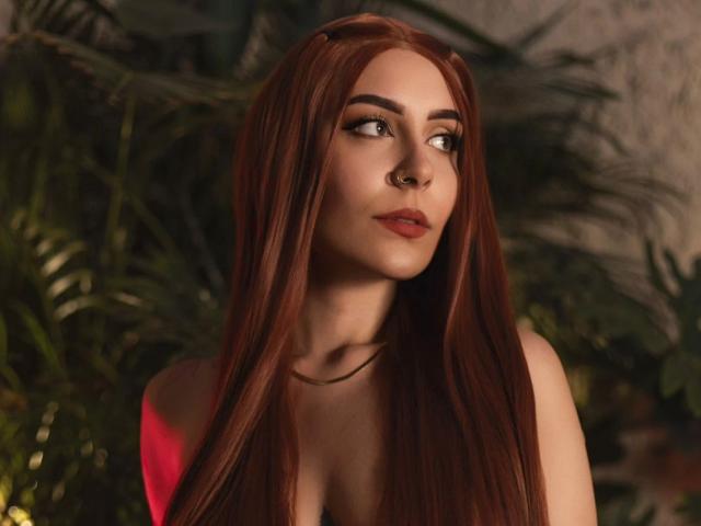 AlesziaRose with long red hair, nose ring, and pink dress, gazing to the side with a slight smile, her tits partially visible, in a softly lit, green background.