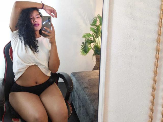 AriaVega with curly black hair, wearing a white tied shirt and black panties, sitting on a chair, taking a mirror selfie, showing her tits and pussy.