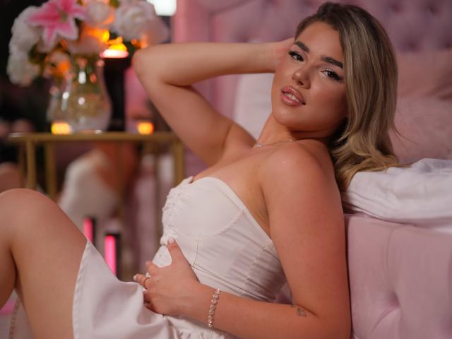 KiaraConti, avec ses cheveux blonds, vêtue d'une robe blanche sans bretelles, tenant ses seins, allongée sur un lit rose, une main derrière la tête, un sourire doux, une lampe rose, des fleurs blanches et un miroir doré en arrière-plan..