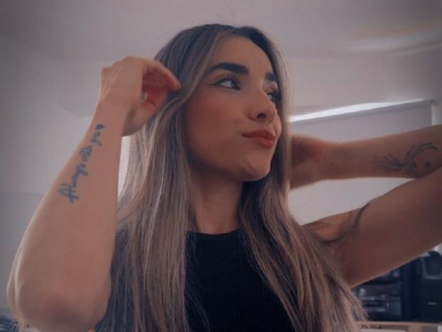 AdharaCooperr with long, straight grayish-blonde hair, pouty red lips, and tattoos on her arm, looking to the side, hands in her hair, wearing a black top.