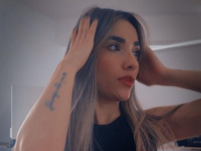 AdharaCooperr has long, blonde hair, red lipstick, and a tattoo on her left arm. She's touching her hair with both hands, looking to the side.