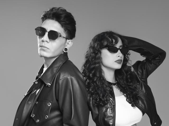 AbrilAndJulian, un hombre con cabello corto y oscuro, chaqueta de cuero y gafas de sol oscuras, está de pie junto a AbrilAndJulian, una mujer con cabello rizado y negro, camisa blanca y chaqueta de cuero..