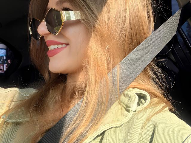 JacqueHailstone, avec ses cheveux blonds illuminés par le soleil, portant des lunettes de soleil aviateur dorées, une veste verte et une ceinture de sécurité, souriant avec du rouge à lèvres, assise dans une voiture..