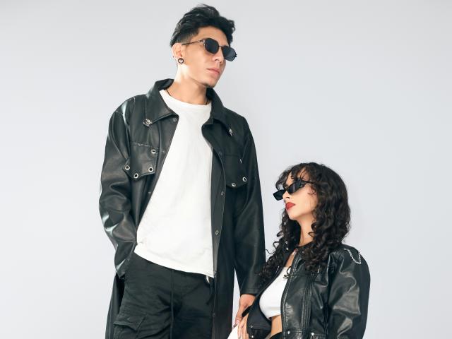 AbrilAndJulian, tall dude with black leather jacket, white tee, black pants, and sunglasses, standing next to AbrilAndJulian, curly-haired chick with black leather jacket, white tee, and glasses.