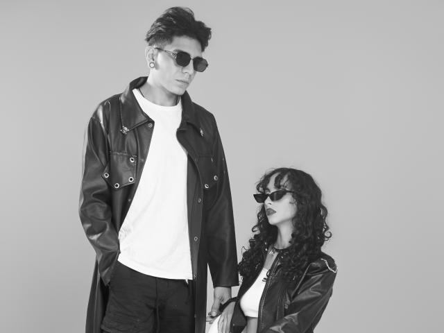 AbrilAndJulian, tall dude with dark leather jacket, white tee, and sunglasses, stands next to AbrilAndJulian, curly-haired chick with leather jacket, dark sunglasses, and white tee.