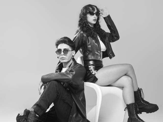 AbrilAndJulian, with dark curly hair and sunglasses, sits on a chair in a leather jacket and shorts, showing her tits and ass. Julian, with dark hair and sunglasses, sits below.