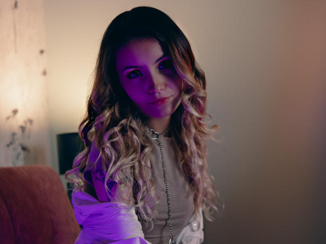 LilaLaurent with long, wavy blonde hair, purple light on her face, white shirt, chain necklace, and a subtle smile, looking directly at the camera.