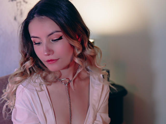LilaLaurent has wavy blonde hair, pink makeup, and a silver chain collar. She's wearing a low-cut white top, looking down with a subtle smile.