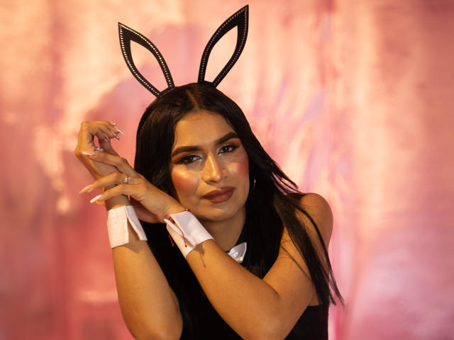 SilvanaCastro with black hair, wearing bunny ears, black dress, white cuffs, and pink makeup, holding her hands near her face, showing off her nails and ring.