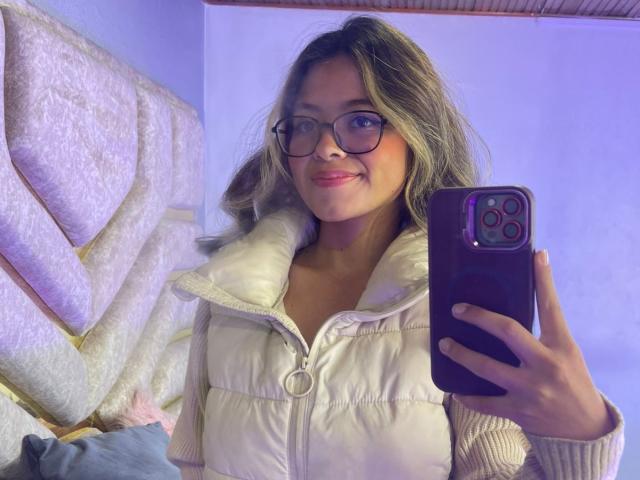 AnittaRouses with glasses, smiling, holding a purple phone, wearing a white puffy vest, and pink lipstick, has blonde wavy hair, and is taking a selfie.