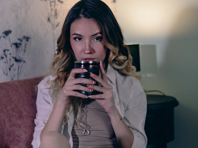 LilaLaurent with blonde curls, holding a black can, wearing a white shirt and beige top, sitting on a pink couch, looking into the camera with serious eyes.