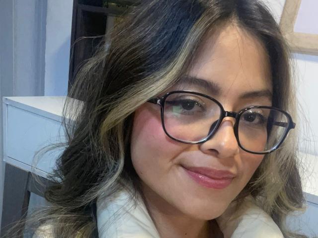 AnittaRouses has medium brown skin, wavy blonde and brown hair, black glasses, pink blush, and a subtle smile. She's wearing a white shirt, with visible pink lips.