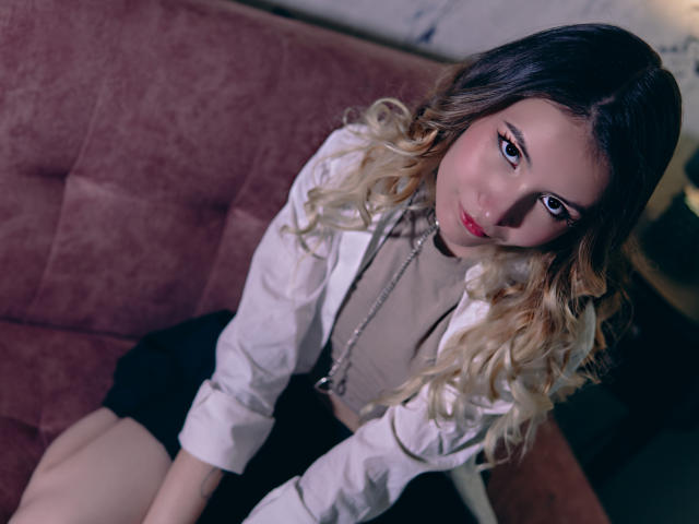 LilaLaurent with blonde curls, wearing a white jacket, sitting on a pink couch, looking up with pink lips and makeup, black skirt, and a neutral top.