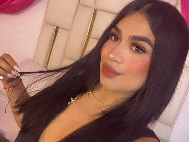 SilvanaCastro with long black hair, holding a red thread, wearing a black top and gold cross necklace, smiling with pink blush, brown eyes, and full lips.