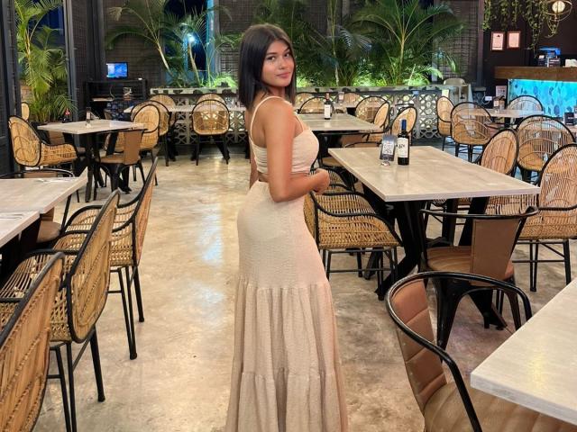 AshlyLee está de pie en un restaurante, vestida con un vestido ajustado color beige transparente que resalta sus glúteos y sus pequeños senos. Tiene el pelo oscuro y una sonrisa encantadora..