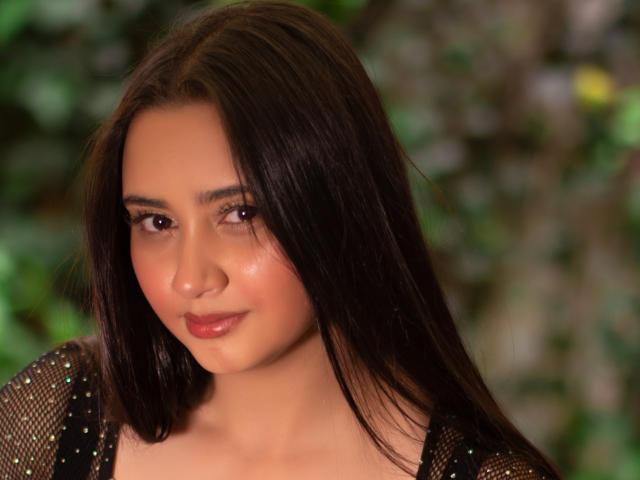 GraceBlue69 with shiny brown hair, subtle makeup, and glossy pink lips, wearing a see-through black top with tiny sparkles, looking down with a gentle smile.