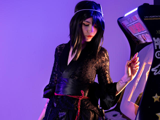 LilaLaurent with black lace kimono, red belt, and black bob, stands by a purple-lit arcade machine, looking down with a gentle, focused expression. Her hand touches the machine.