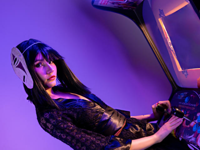 LilaLaurent with black hair, wearing a white mask, black lace top, and leather vest, holding a game machine, illuminated by purple light, looking serious.