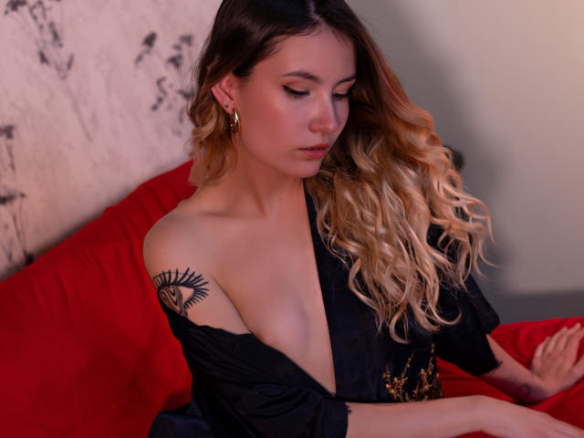 LilaLaurent with wavy blonde hair, black off-shoulder top, gold earrings, sun tattoo on left shoulder, sitting on red bed, looking down, soft lighting, subtle makeup.