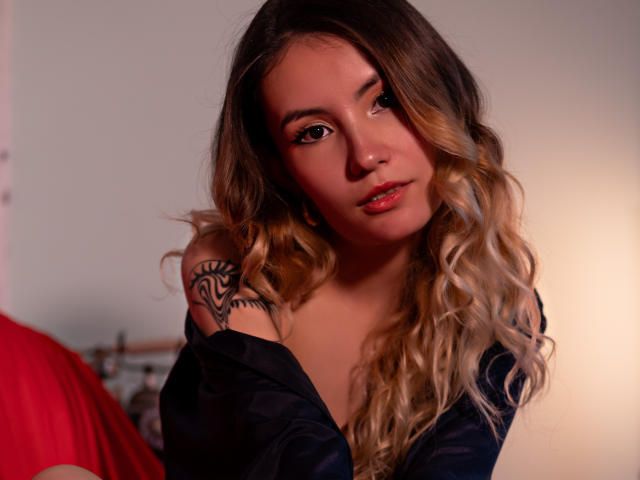 LilaLaurent with long, wavy blonde hair, dark eye makeup, glossy pink lips, black off-shoulder top, tattoo on right shoulder, red fabric in background, soft lighting, subtle smile.