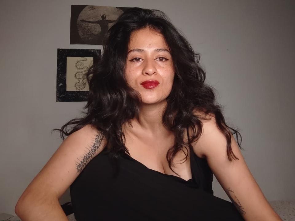 Una succube con capelli neri ondulati, rossetto rosso e un tatuaggio sul braccio destro, che indossa una canotta nera, guarda direttamente la fotocamera con un leggero sbuffo..