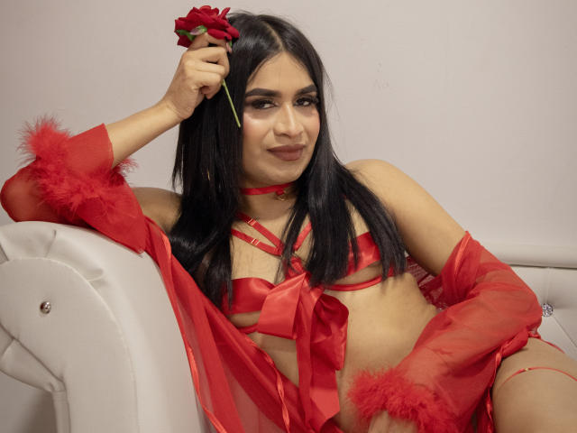 SilvanaCastro with long black hair, red lingerie, and red feathered arm cuffs, holding a red rose to her head, sitting on a white couch, smiling. Her tits are visible.